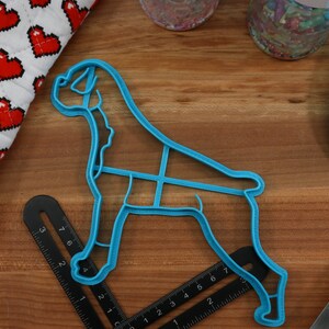 May include: A blue plastic cookie cutter shaped like a boxer dog. The cutter is on a wooden surface with a ruler and a blue spatula.