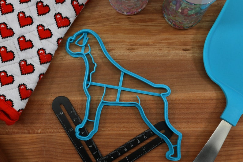 May include: A blue plastic cookie cutter shaped like a boxer dog. The cutter is on a wooden surface with a ruler and a blue spatula.