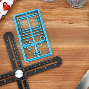 May include: A blue plastic cookie cutter shaped like a vintage handheld video game console. The cutter is on a wooden surface with a black ruler and a blue spatula.