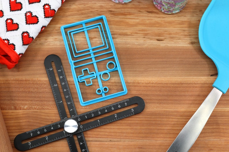 May include: A blue plastic cookie cutter shaped like a vintage handheld video game console. The cutter is on a wooden surface with a black ruler and a blue spatula.