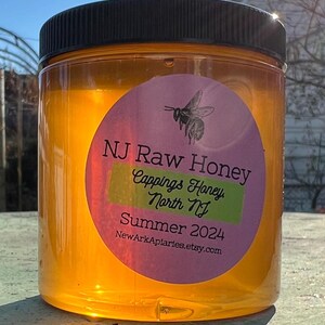 May include: A glass jar filled with honey, labeled "NJ Raw Honey, Capping's Honey, North NJ, Summer 2024, NewarkAplarles.etsy.com".