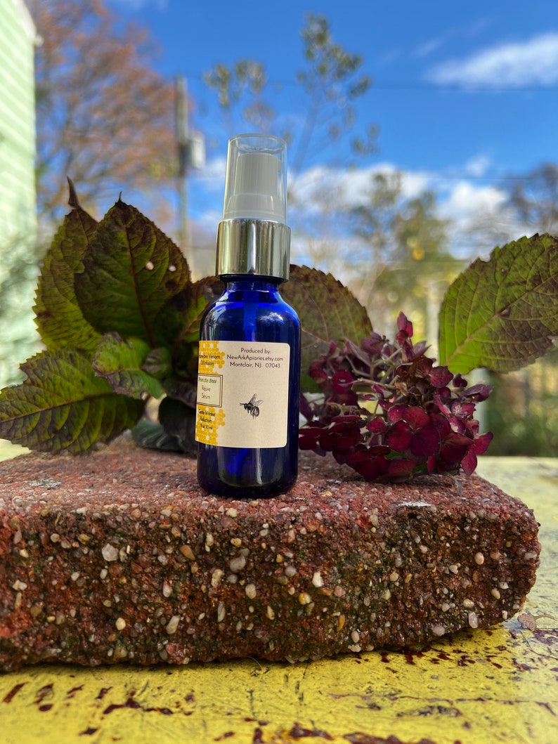 May include: A blue glass bottle with a silver cap and a white spray nozzle. The bottle is labeled with the text "Produced by: NewApiapis.etsy.com  Montclair, NJ 07043". A small bee illustration is on the label.