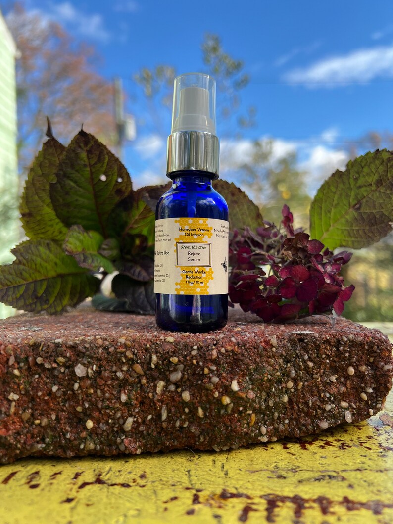 May include: A small blue glass bottle with a white spray top. The bottle is labeled "Honeybee Venom Oil Infusion" and "Gentle Wrinkle Reduction". The bottle is sitting on a red brick.