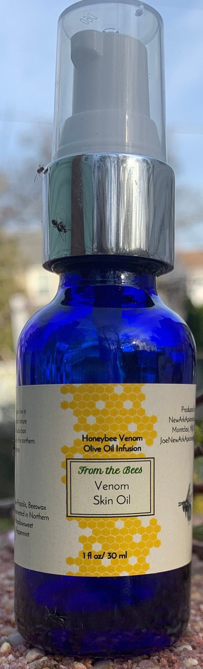 May include: A blue glass bottle with a silver pump top and a white label. The label has a honeycomb pattern and the text "Honeybee Venom Olive Oil Infusion From the Bees Venom Skin Oil 1 fl oz/30 ml".