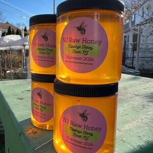 May include: Three glass jars of honey with black lids. The jars are labeled "NJ Raw Honey" with a pink and green circle sticker. The text on the sticker reads "Cappings Honey North NJ Summer 2024" and a website address. The jars are sitting on a wooden surface.