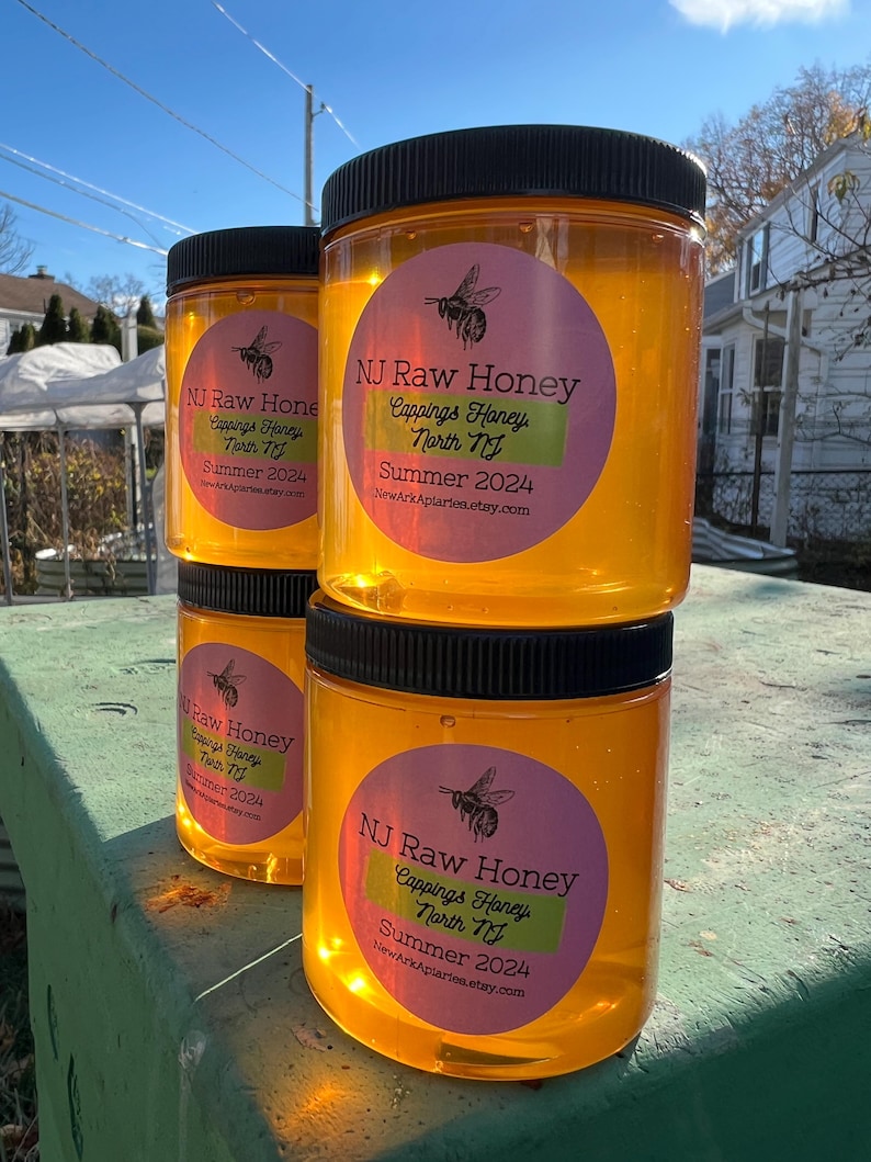 May include: Three glass jars of honey with black lids. The jars are labeled "NJ Raw Honey" with a pink and green circle sticker. The text on the sticker reads "Cappings Honey North NJ Summer 2024" and a website address. The jars are sitting on a wooden surface.