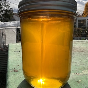 May include: A clear glass jar filled with a golden yellow liquid, likely honey, with a silver lid. The jar is sitting on a green surface.