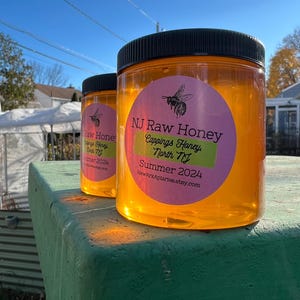 May include: Two glass jars of honey with black lids and pink and green labels. The labels say "NJ Raw Honey Cappings Honey North NJ Summer 2024 NewArkAplaries.etsy.com".