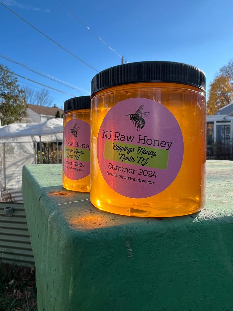 May include: Two glass jars of honey with black lids and pink and green labels. The labels say "NJ Raw Honey Cappings Honey North NJ Summer 2024 NewArkAplaries.etsy.com".