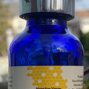 May include: A blue glass bottle with a silver pump top and a white label. The label has a honeycomb pattern and the text "Honeybee Venom Olive Oil Infusion From the Bees Venom Skin Oil 1 fl oz/30 ml".