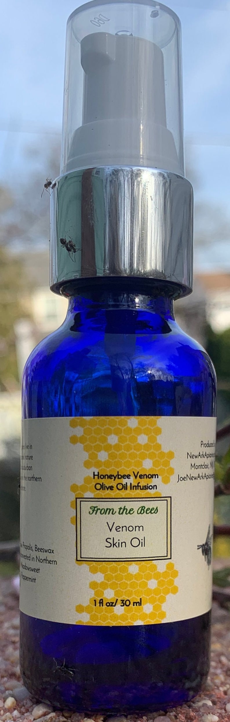 May include: A blue glass bottle with a silver pump top and a white label. The label has a honeycomb pattern and the text "Honeybee Venom Olive Oil Infusion From the Bees Venom Skin Oil 1 fl oz/30 ml".