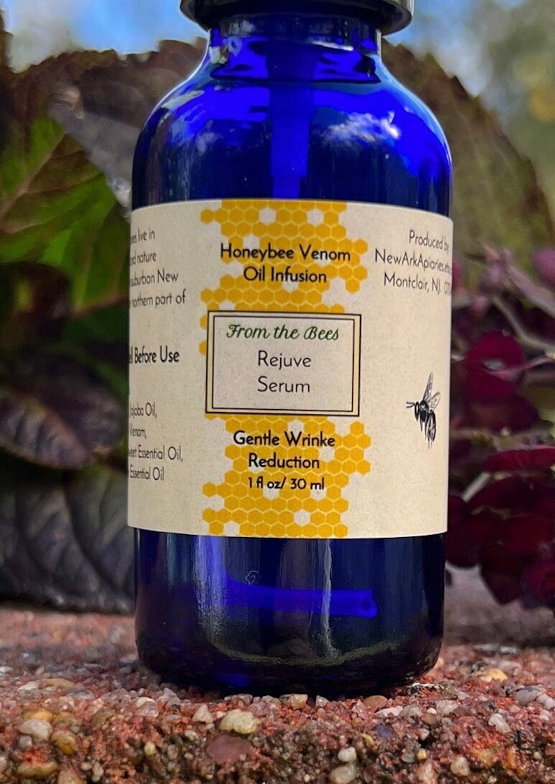 May include: A blue glass bottle with a label that reads "Honeybee Venom Oil Infusion", "From the Bees Rejuve Serum", "Gentle Wrinkle Reduction", and "1 fl oz/30 ml". The label has a honeycomb pattern and a small bee illustration.