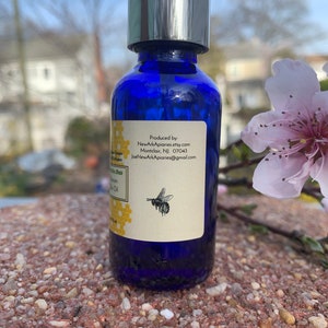 May include: A blue glass bottle with a silver pump top and a white label. The label has text that reads "Produced by: NewArkApiaries.etsy.com Montclair, NJ 07043 JoeNewArkApiaries@gmail.com" and a small image of a bee. The bottle is sitting on a brown, rocky surface.
