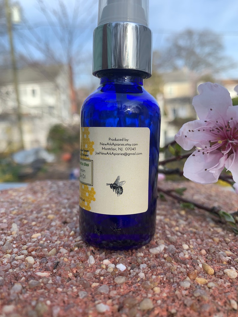 May include: A blue glass bottle with a silver pump top and a white label. The label has text that reads "Produced by: NewArkApiaries.etsy.com Montclair, NJ 07043 JoeNewArkApiaries@gmail.com" and a small image of a bee. The bottle is sitting on a brown, rocky surface.