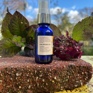 May include: A blue glass bottle with a silver spray nozzle and a white label. The label has text that reads "Our honeybees live in backyards and nature preserves in suburban New Jersey in the northern part of the store. Shake Well Before Use. Ingredients: Argan Oil, Jojoba Oil, Honeybee Venom, Meadowsweet Essential Oil, Immortelle Essential Oil."