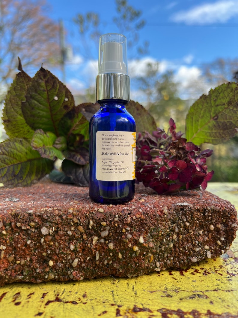 May include: A blue glass bottle with a silver spray nozzle and a white label. The label has text that reads "Our honeybees live in backyards and nature preserves in suburban New Jersey in the northern part of the store. Shake Well Before Use. Ingredients: Argan Oil, Jojoba Oil, Honeybee Venom, Meadowsweet Essential Oil, Immortelle Essential Oil."