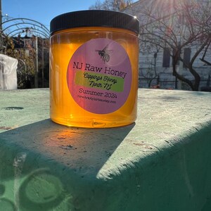 May include: A clear glass jar of honey with a label that reads "NJ Raw Honey, Cappings Honey, North NJ, Summer 2024, NewArkApiaries.etsy.com". The jar is sitting on a green surface.