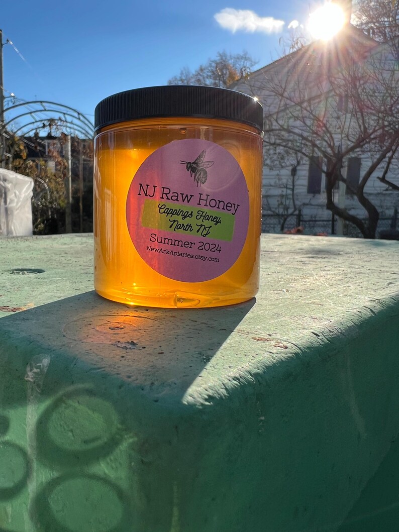 May include: A clear glass jar of honey with a label that reads "NJ Raw Honey, Cappings Honey, North NJ, Summer 2024, NewArkApiaries.etsy.com". The jar is sitting on a green surface.