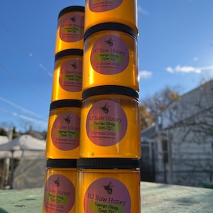 May include: A stack of six amber glass jars filled with honey. Each jar has a black lid and a pink and green label that reads "NJ Raw Honey Cappings Honey North NJ Summer 2024".