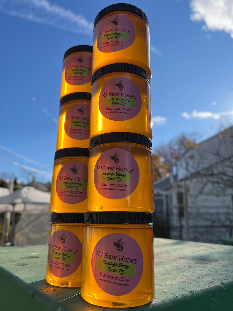 May include: A stack of six amber glass jars filled with honey. Each jar has a black lid and a pink and green label that reads "NJ Raw Honey Cappings Honey North NJ Summer 2024".