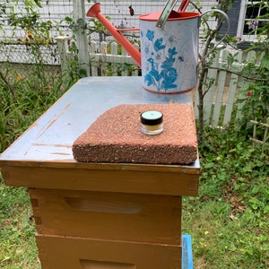 May include: A wooden beehive with three boxes stacked on top of each other. The top box has a metal lid with a small, round, white jar on top of a red brick. A red watering can with a blue and white floral pattern is behind the beehive.