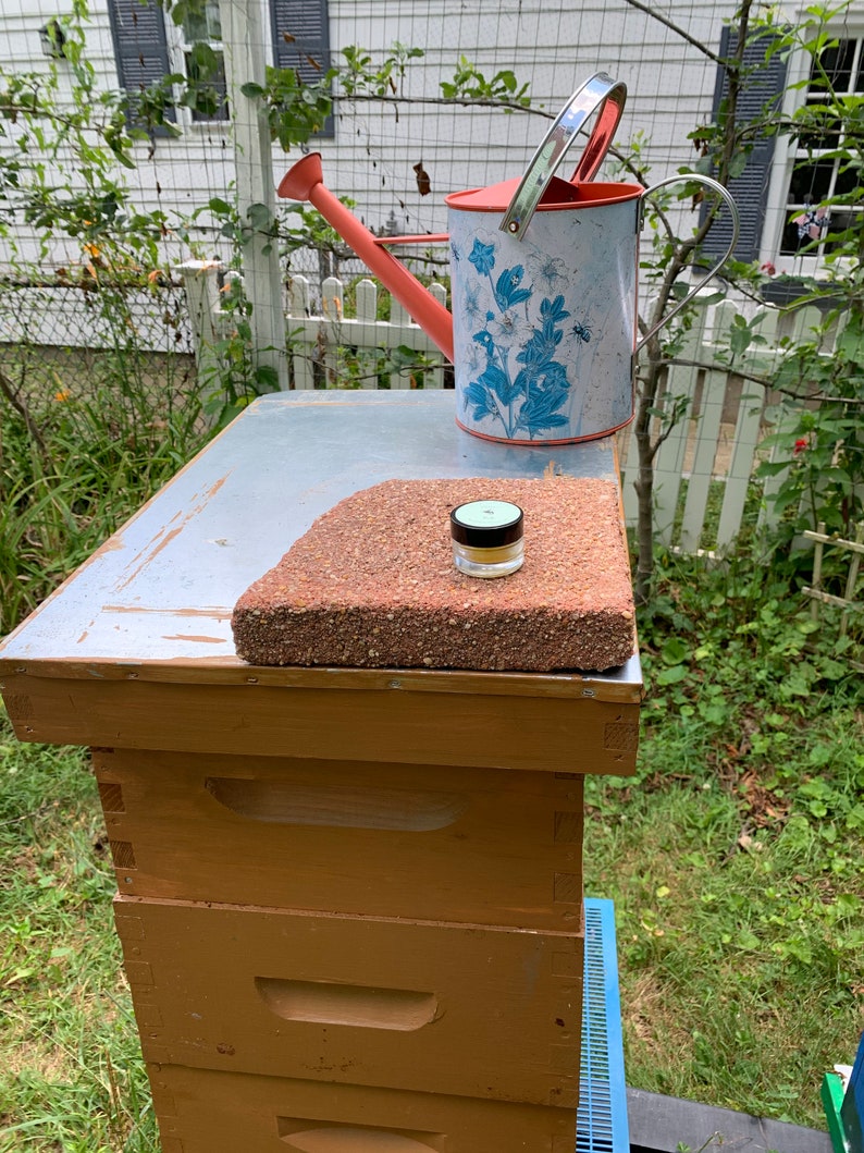 May include: A wooden beehive with three boxes stacked on top of each other. The top box has a metal lid with a small, round, white jar on top of a red brick. A red watering can with a blue and white floral pattern is behind the beehive.