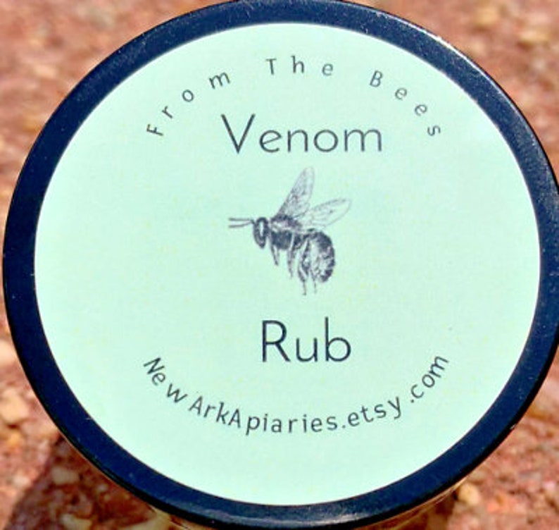 May include: A round jar with a black lid and a green label. The label says "From The Bees Venom Rub New ArkApiaries.etsy.com". A black and white illustration of a bee is on the label.