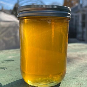 May include: A glass jar filled with golden honey, with a silver lid. The jar is sitting on a green surface.