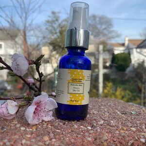 May include: A blue glass bottle with a silver pump top and a white label. The label says "Honeybee Venom Olive Oil Infusion" and "From the Best Venom Skin Oil". The bottle is sitting on a brown, textured surface.