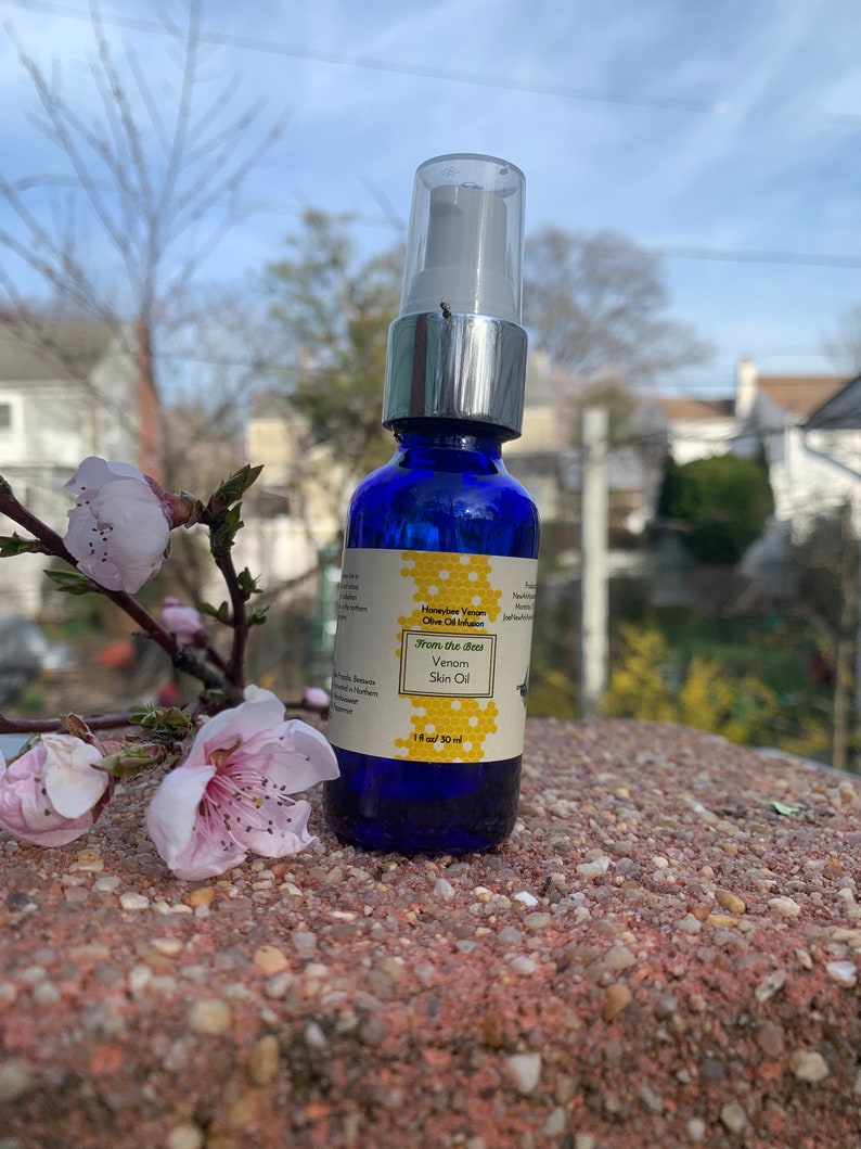 May include: A blue glass bottle with a silver pump top and a white label. The label says "Honeybee Venom Olive Oil Infusion" and "From the Best Venom Skin Oil". The bottle is sitting on a brown, textured surface.