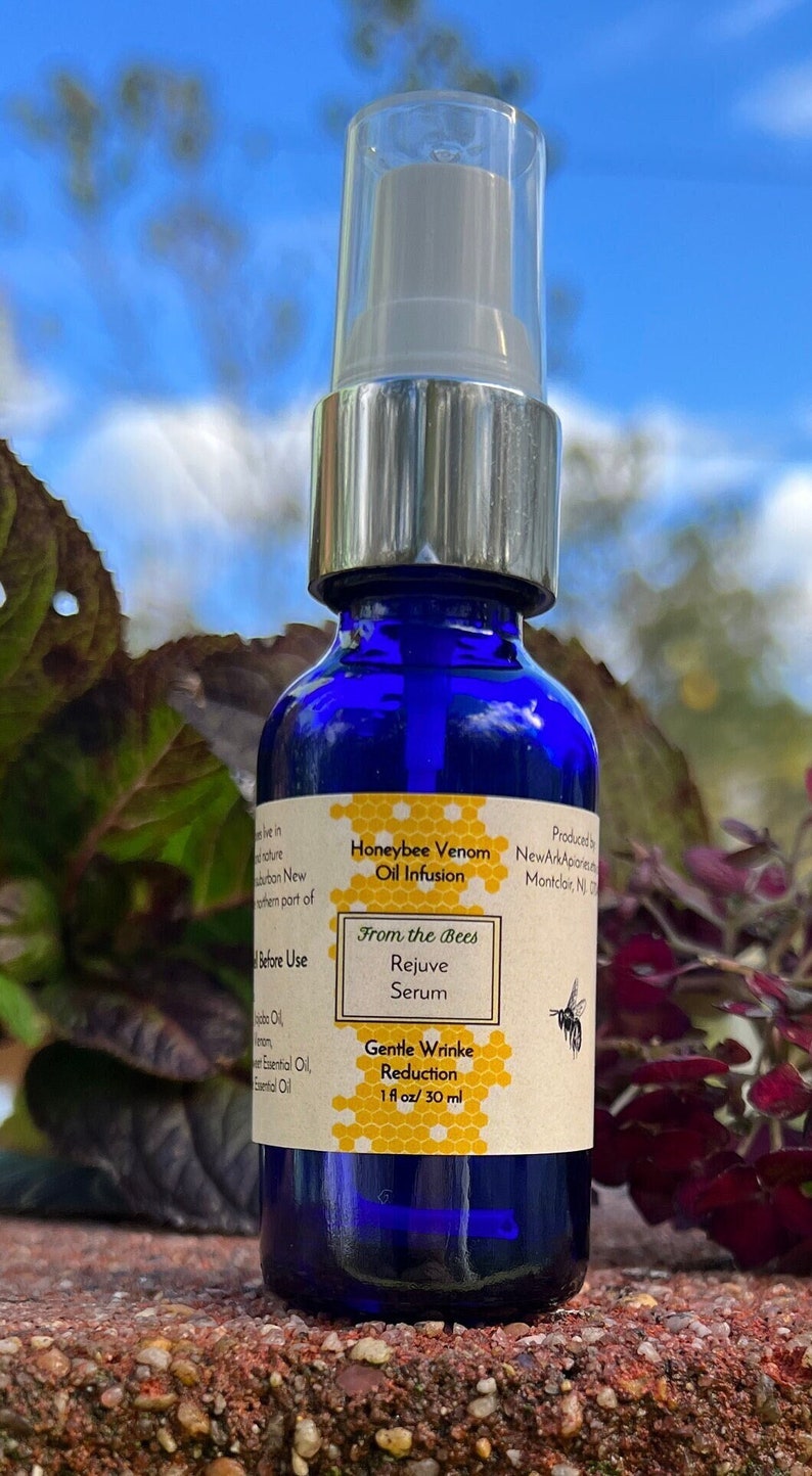 May include: A blue glass bottle with a silver cap and a label that reads "Honeybee Venom Oil Infusion From the Bees Rejuve Serum Gentle Wrinkle Reduction 1 fl oz/30 ml". The bottle is sitting on a brown surface with green plants in the background.