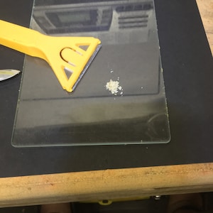 May include: A yellow plastic scraper with a triangular blade sits on a clear glass surface. A small pile of white powder is on the glass surface.