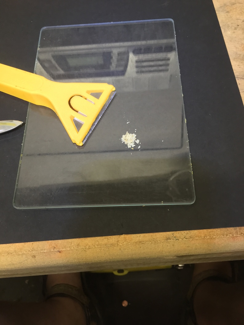 May include: A yellow plastic scraper with a triangular blade sits on a clear glass surface. A small pile of white powder is on the glass surface.