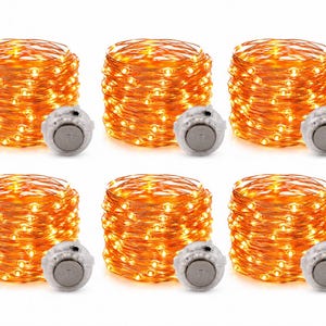 May include: A set of six coiled copper wire string lights, each featuring small warm yellow LED bulbs and a circular silver battery pack with a small switch, arranged in two rows of three against a plain white background.