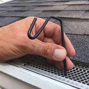 May include: A black metal gutter guard hook is being held by a hand. The hook is designed to secure gutter guards to the gutter.