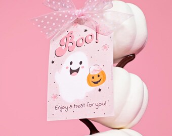 Pode incluir: Uma etiqueta de presente retangular com um fantasma branco segurando uma abóbora laranja, com o texto "Boo!" e "Enjoy a treat for you!" em rosa. A etiqueta é finalizada com um laço de fita rosa transparente com bolinhas e repousa sobre abóboras brancas.