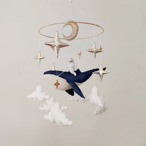 May include: A gold metal mobile with a blue whale, a white astronaut, and gold stars and a crescent moon hanging from it. White felt clouds are also hanging from the mobile.