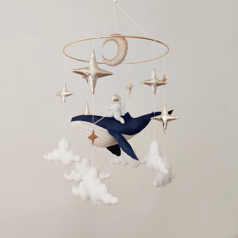 May include: A gold metal mobile with a blue whale, a white astronaut, and gold stars and a crescent moon hanging from it. White felt clouds are also hanging from the mobile.