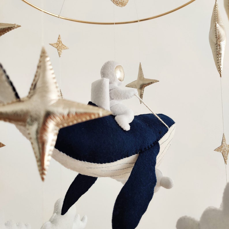 May include: A white astronaut figurine riding a blue whale mobile with gold glitter stars hanging from a gold metal ring.