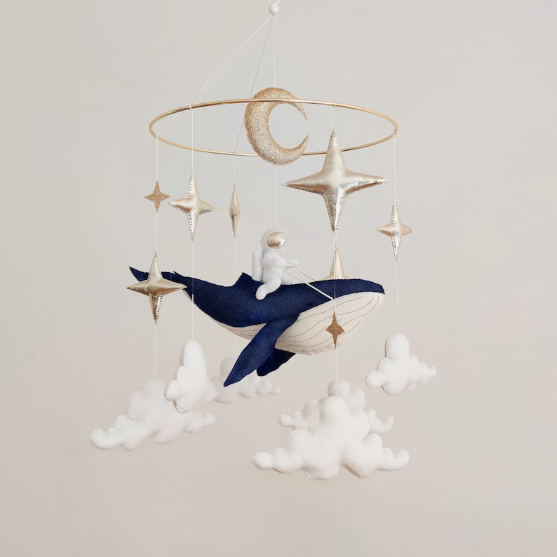 May include: A blue whale mobile with a gold astronaut, stars, and clouds. The whale is made of felt and has a white stitched line down its back. The astronaut is holding a rope attached to the whale.