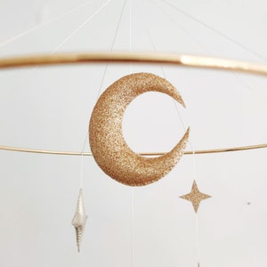 May include: A gold glitter crescent moon and a gold glitter star hang from a gold metal hoop mobile.