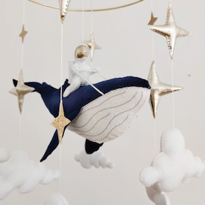 May include: A blue whale mobile with a white astronaut riding on top. The whale is surrounded by white clouds and gold stars.