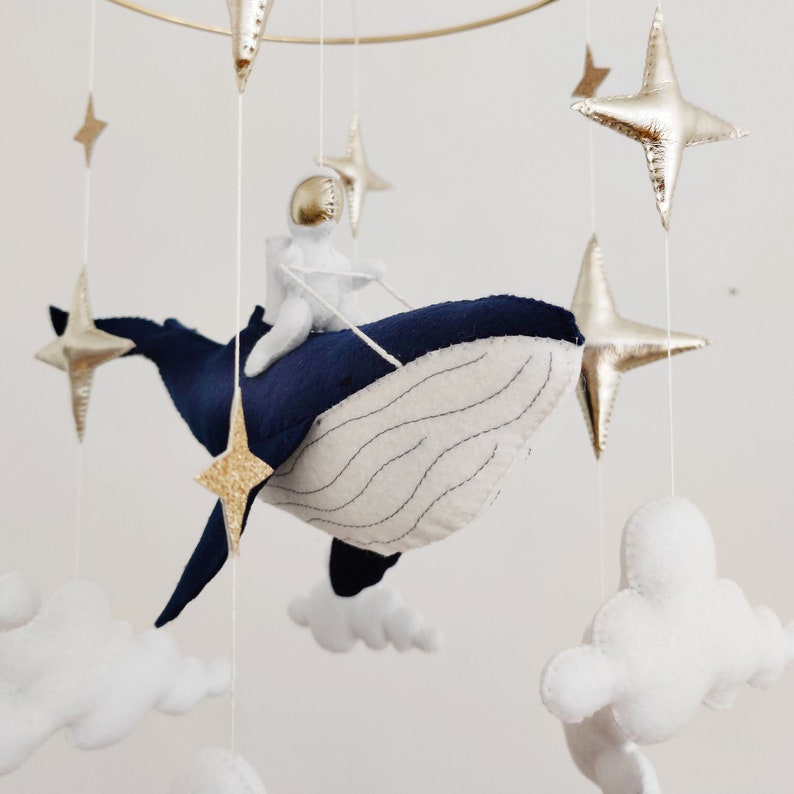 May include: A blue whale mobile with a white astronaut riding on top. The whale is surrounded by white clouds and gold stars.