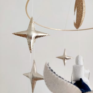 May include: A gold and white mobile with a crescent moon, stars, and a cloud. The mobile is hanging from a gold hoop.