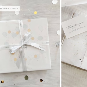 May include: White gift box with a white ribbon and gold confetti. The ribbon has the text "ZOZOTOYS" printed on it. The box is sitting on a white wooden surface. A thank you card is included with the gift.