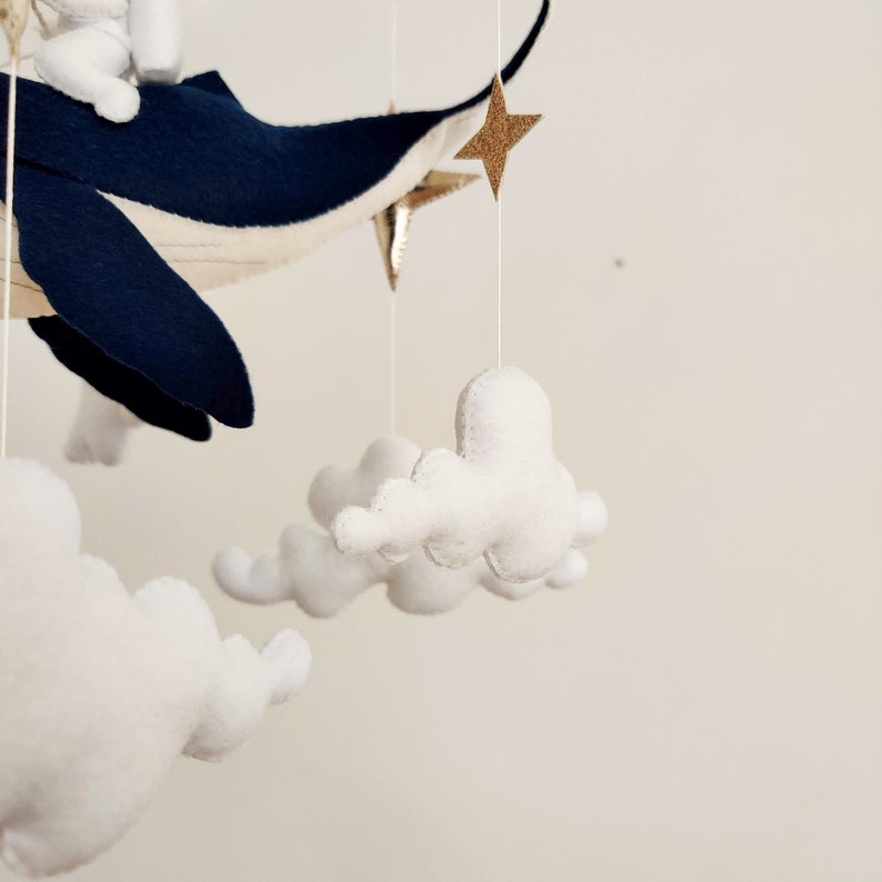 May include: A white felt whale mobile with white felt clouds and gold glitter stars hanging from it. The whale is flying through the air.