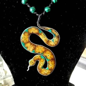 Può includere: Una collana con un ciondolo a forma di serpente in pietra di malachite verde. Il serpente è in metallo e presenta un motivo dorato e verde.