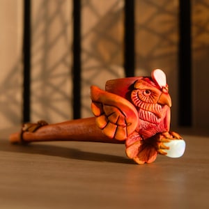 May include: A hand-crafted ceramic owl pipe with a red and orange glaze. The owl is holding a small, white, round object in its talons.