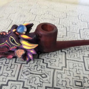 May include: A wooden smoking pipe with a dragon head design. The dragon is painted in shades of purple, yellow, and black with a blue glass eye. The pipe is made of dark wood and has a long, curved stem.