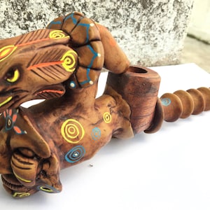May include: A wooden pipe with a carved eagle head and a human face. The pipe is decorated with colorful paint and swirls. The pipe is made of wood and has a long, thin stem.